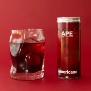 Image of a can of Americano and a glass containing the drink next to it. The dominant color of the image is red, present in the can's graphics, the color of the drink, and the background.