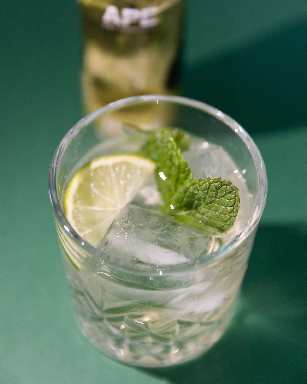 immagine di un bicchiere fotografato dall'alto contenente l'aperitivo Hugo, ghiaccio e una fettina di lime. Lo sfondo è verde foresta.