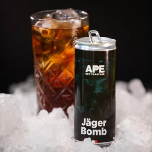 Image of a can of Jäger Bomb in the foreground and a glass containing the cocktail in the background. The can and the glass are placed on top of crushed ice. The background and the can's graphics are black.