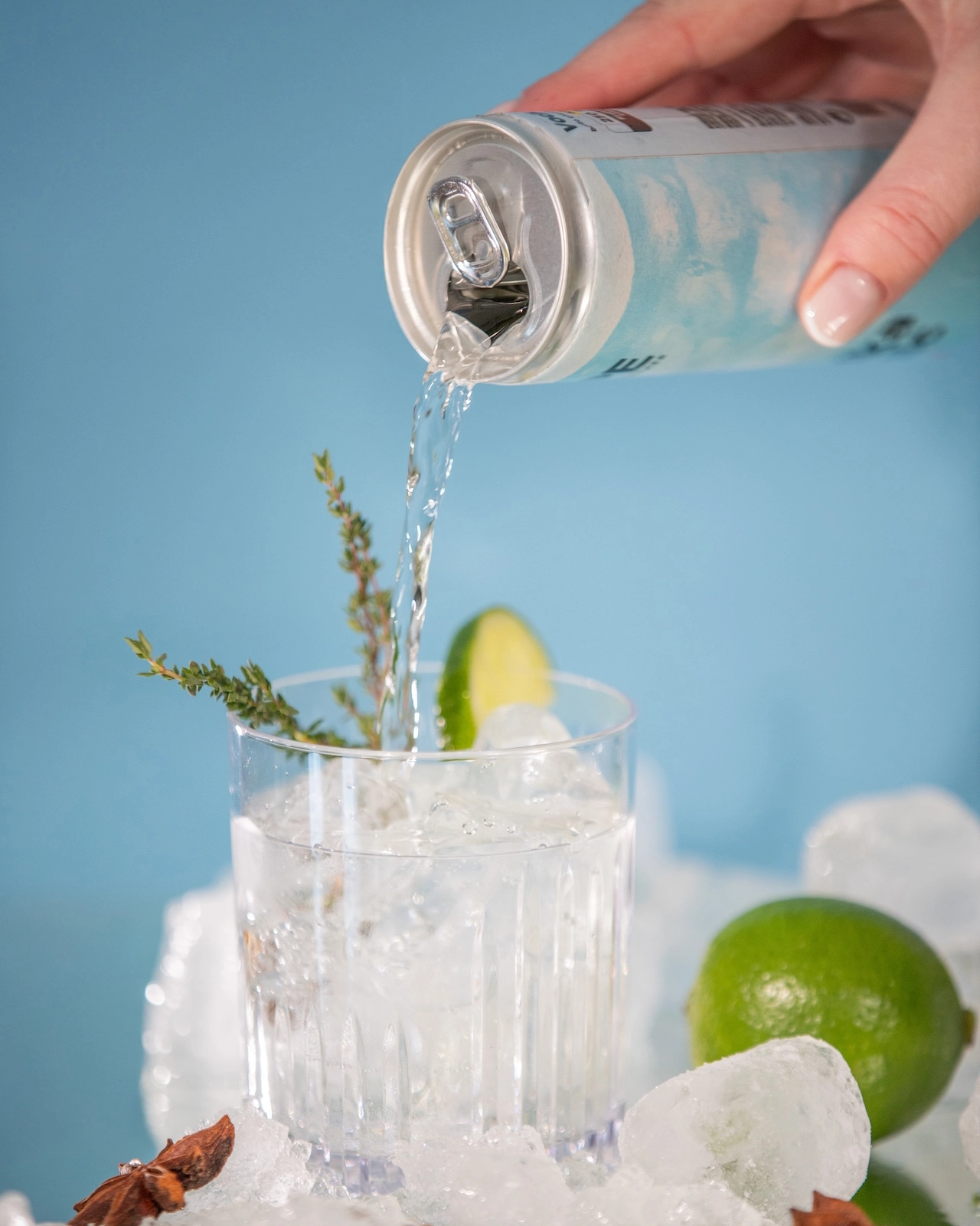 immagine di una mano che versa il contenuto di una lattina di vodka tonic su un bicchiere. Nel bicchiere è presente ghiaccio, un rametto di contorno e una fettina di lime. Lo sfondo è azzurro