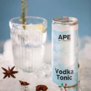 Image of a can of vodka tonic in the foreground and a glass containing the cocktail in the background. There is crushed ice and star anise as compositional elements in the image. The background is blue.